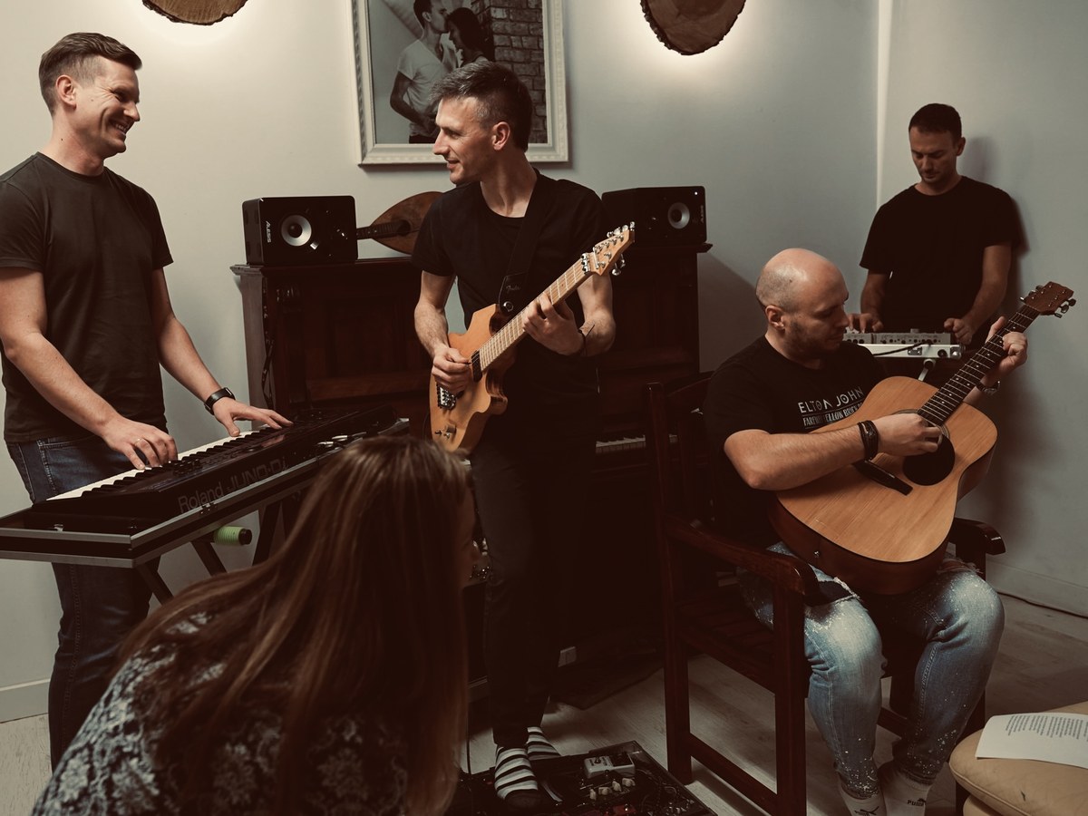 Warsztaty Człowiek-Orkiestra — uczestnicy grają razem na gitarach, keyboardzie i sprzęcie muzycznym, uśmiechnięci w trakcie wspólnej sesji